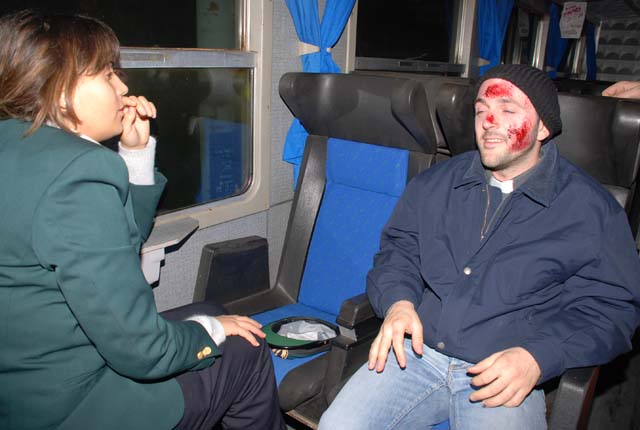 Di spalle una ragazza vestita con la divisa delle Ferrovie dello Stato e in secondo piano un passeggero con delle ferite evidenti